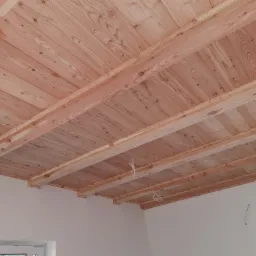 Drewniany sufit z belek, wykonany z jasnego drewna, widoczny z perspektywy wnętrza. Elementy wykończeniowe przy ścianach. Naturalne odcienie drewna.