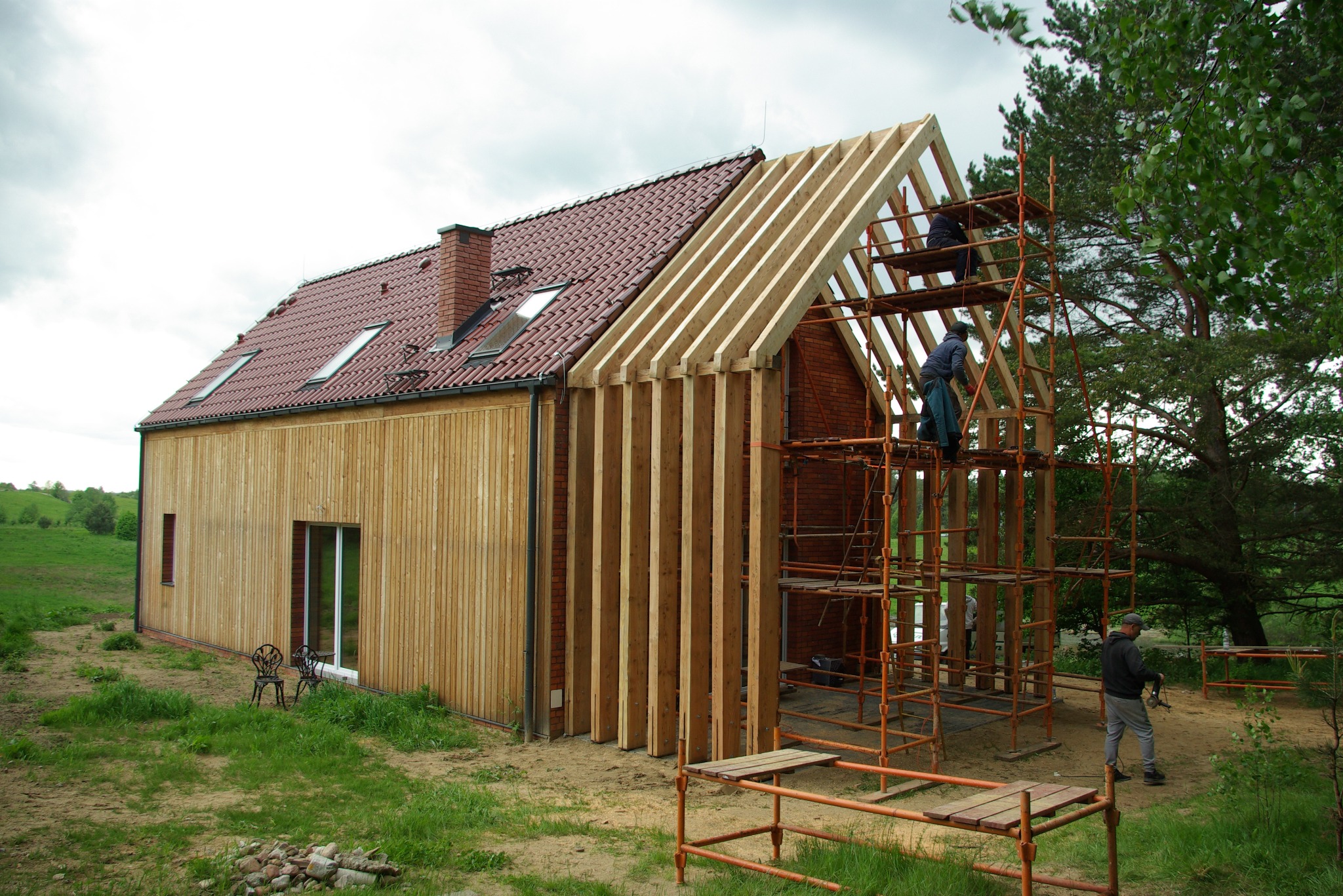 Budowa domu w toku: drewniana elewacja, czerwona dachówka, robotnicy na rusztowaniu montują konstrukcję dachu. Widok na budynek w otoczeniu zieleni.