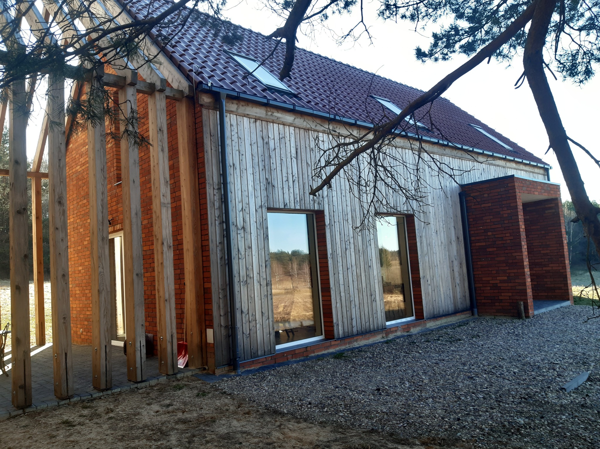 Nowoczesny dom z drewnianą elewacją i ceglanymi elementami, widoczny dach z oknami połaciowymi. Ujęcie z zewnątrz, w naturalnym otoczeniu.