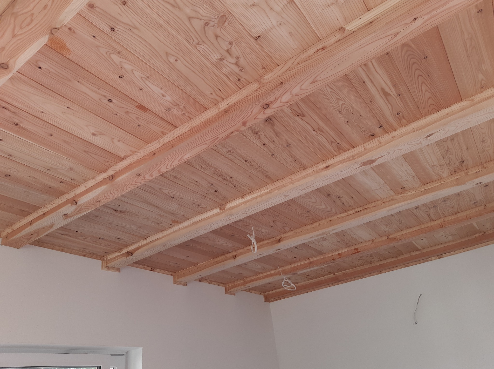 Drewniany sufit z belek, wykonany z jasnego drewna, widoczny z perspektywy wnętrza. Elementy wykończeniowe przy ścianach. Naturalne odcienie drewna.