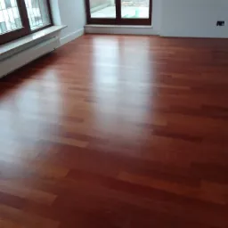 Błyszcząca, nowa podłoga z paneli drewnianych w jasnym, pustym pokoju z dużymi oknami i grzejnikiem pod parapetem.