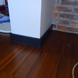 Szlif-Pol Mirosław Lis - Błyszcząca, ciemna drewniana podłoga z widocznym fragmentem białej kolumny z czarną podstawą oraz fragmentem ściany z czerwonej cegły, w tle widoczne przedmioty i fragment podłogi przykryty...