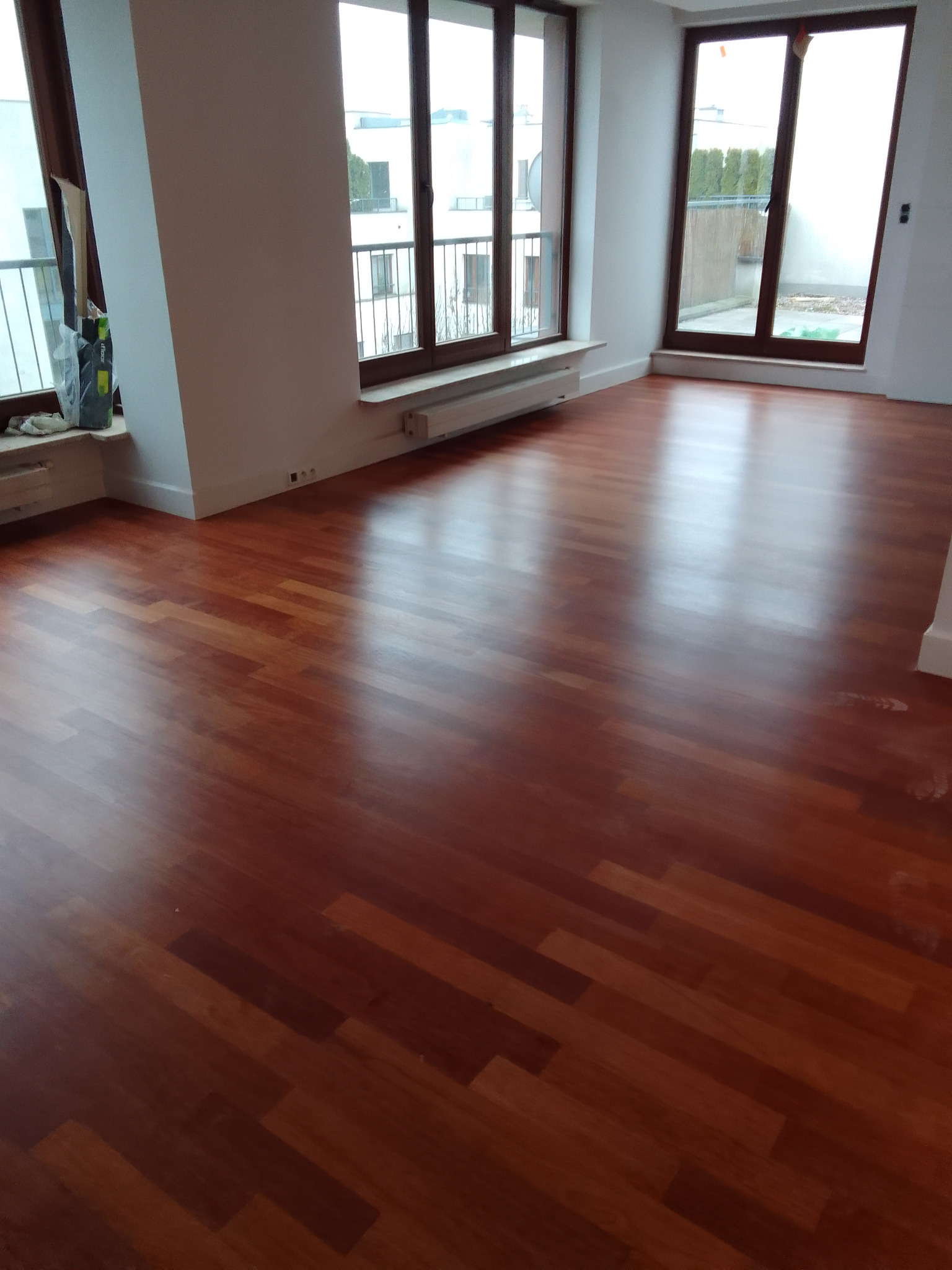 Polakierowana, parkietowa podłoga w odcieniach brązu, ułożona w jodełkę, w jasnym pokoju z dużymi oknami w Warszawie.