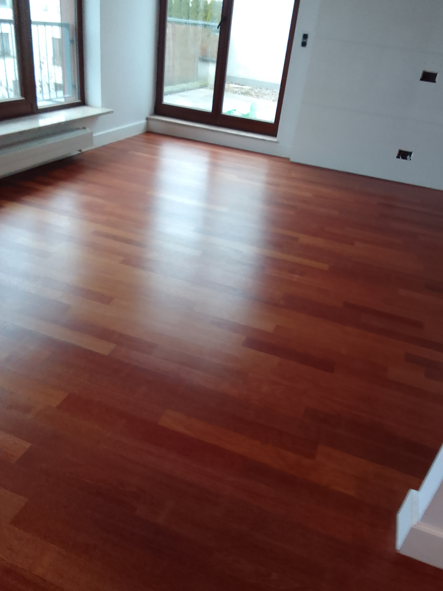 Błyszcząca, świeżo położona podłoga z ciemnego drewna w pustym pokoju z oknami i białymi ścianami, widoczne niedokończone gniazdka elektryczne.