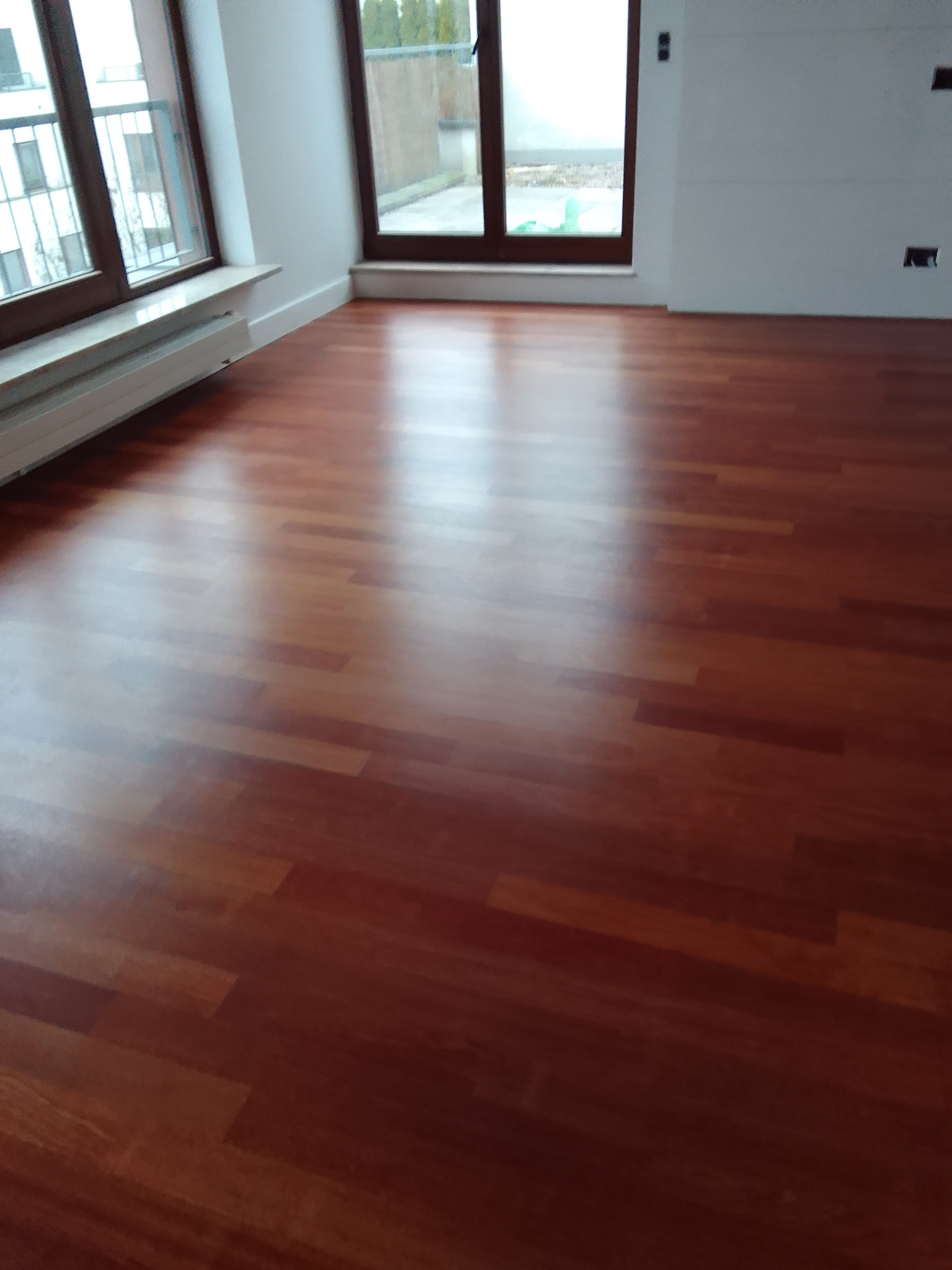 Błyszcząca, nowa podłoga z paneli drewnianych w jasnym, pustym pokoju z dużymi oknami i grzejnikiem pod parapetem.