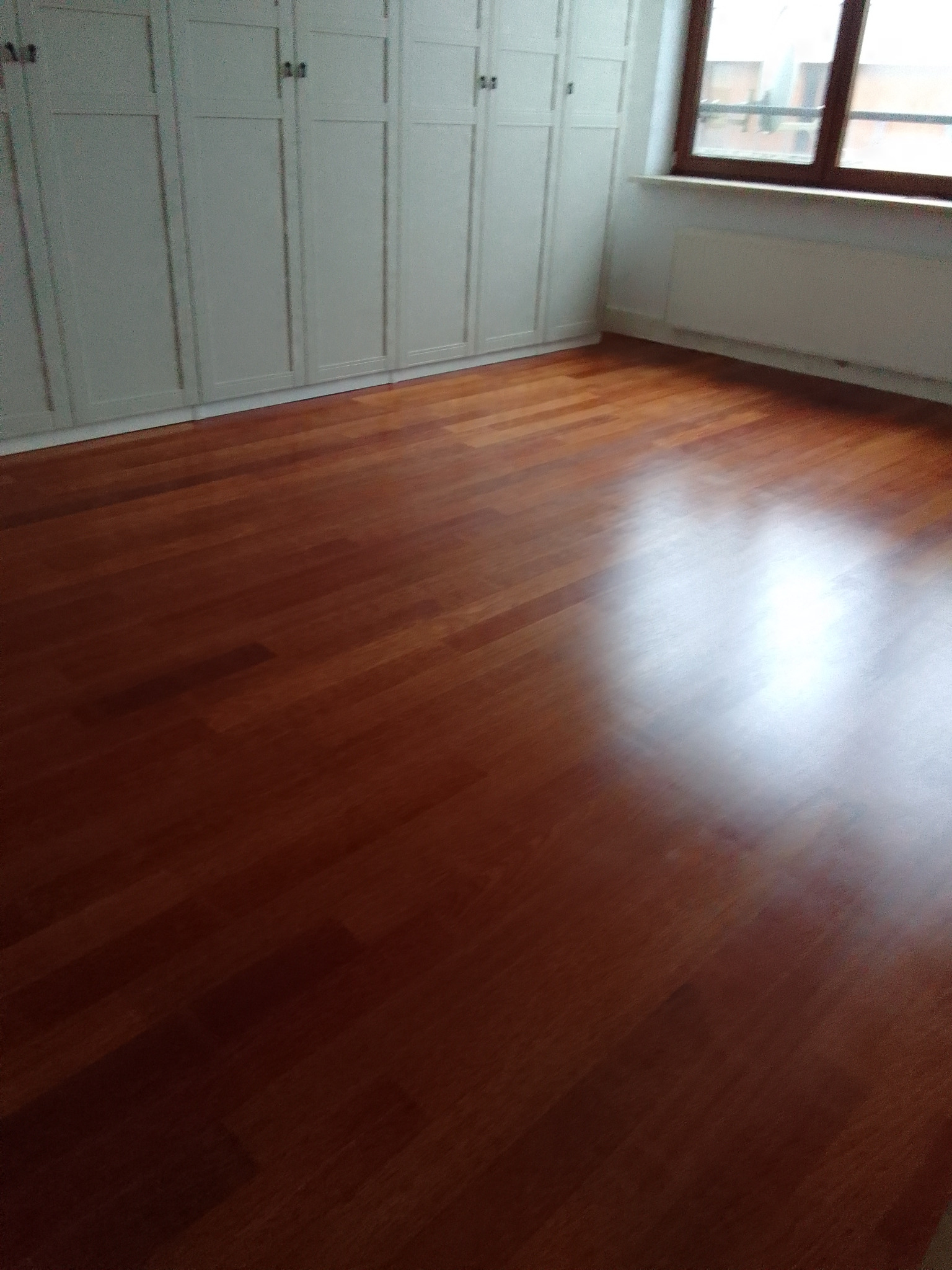 Wypolerowana, ciemna podłoga z paneli drewnianych w jasnym pokoju z białymi szafami w zabudowie i oknem, widoczny fragment grzejnika.