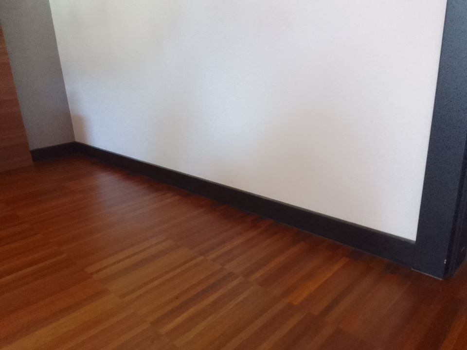 Parkiet z egzotycznego drewna ułożony w jodełkę, przy białej ścianie z czarną listwą przypodłogową i czarną ościeżnicą drzwiową.