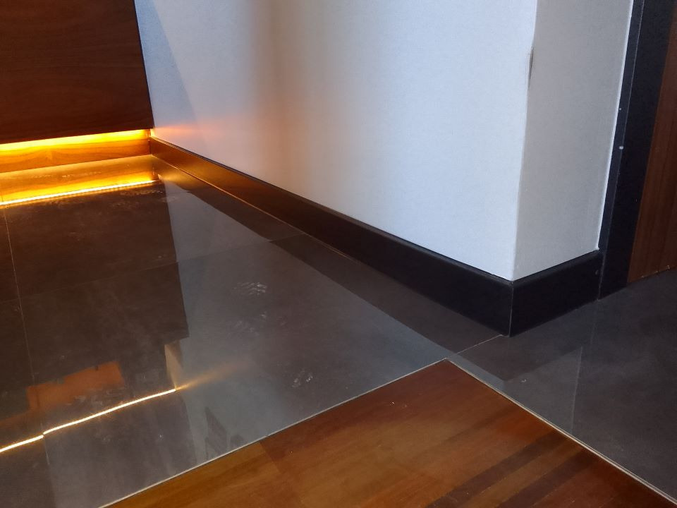 Szare płytki podłogowe z drewnianą wstawką, oświetlenie LED przy ścianie i pod drzwiami, ciemne listwy przypodłogowe.