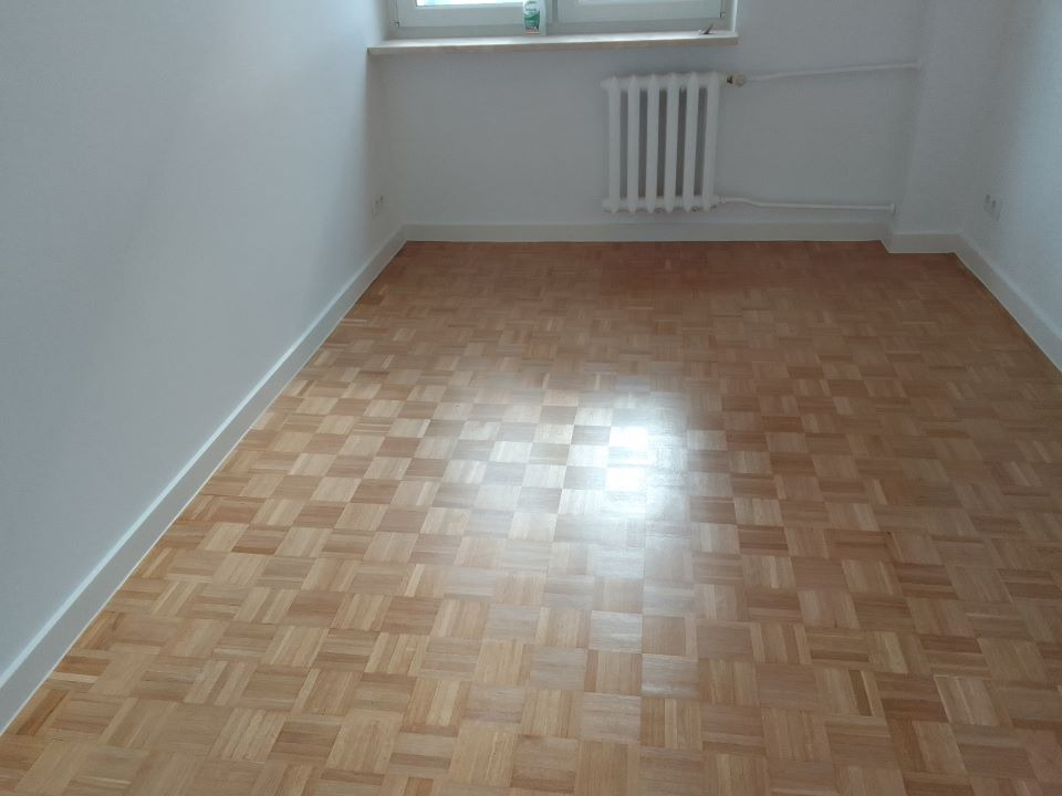 Parkiet ułożony w szachownicę w pustym pokoju z białymi ścianami i grzejnikiem pod oknem.