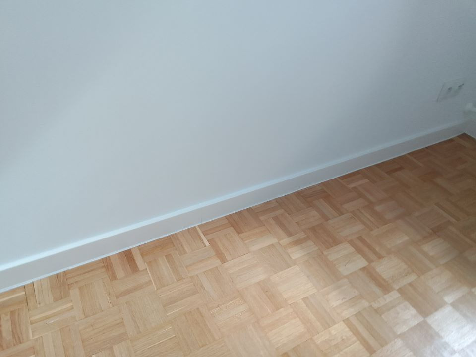 Parkiet ułożony w jodełkę na podłodze, widoczna biała listwa przypodłogowa i fragment białej ściany z gniazdkiem elektrycznym.