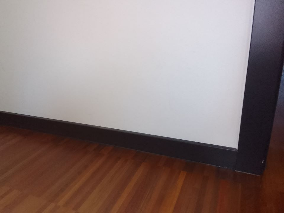 Fragment wnętrza: brązowa drewniana podłoga, biała ściana z ciemną listwą przypodłogową i ciemna rama drzwiowa w prawym rogu.