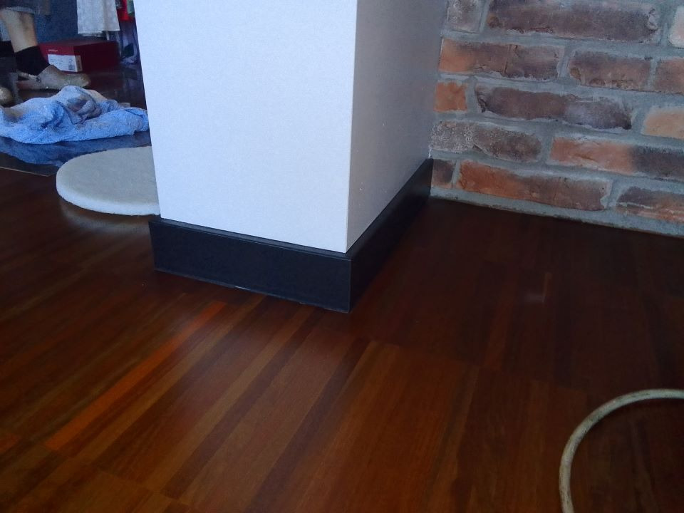 Błyszcząca, ciemna drewniana podłoga z widocznym fragmentem białej kolumny z czarną podstawą oraz fragmentem ściany z czerwonej cegły, w tle widoczne przedmioty i fragment podłogi przykryty...