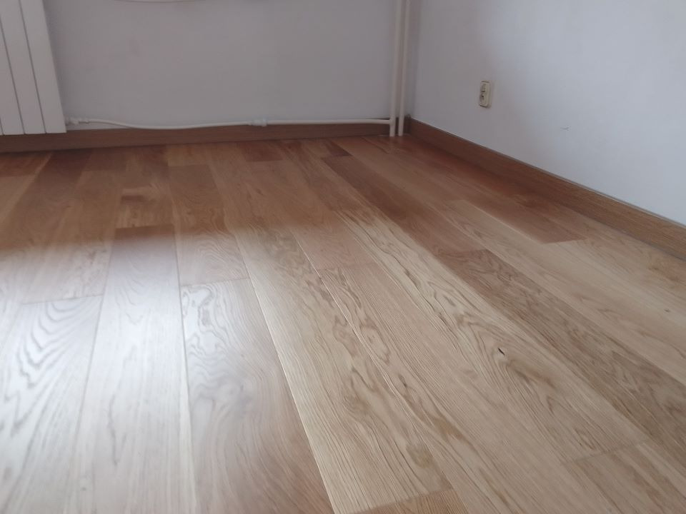 Jasna, drewniana podłoga ułożona w pokoju z białymi ścianami i grzejnikiem, widoczna listwa przypodłogowa.