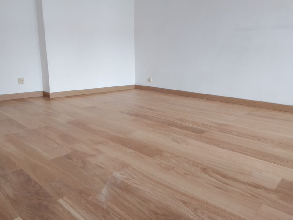 Jasna, świeżo położona podłoga z paneli drewnianych w pustym pokoju z białymi ścianami i listwami przypodłogowymi.