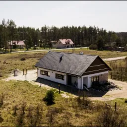 Jednopiętrowy dom z ciemnym dachem i białymi ścianami, widok z góry, otoczony lasem i polami, z widocznym logo firmy na górze zdjęcia.