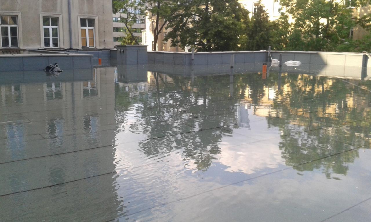 Płaski dach pokryty wodą po deszczu, odbijający budynki i drzewa w kałużach, widoczne elementy konstrukcyjne dachu i fragment elewacji budynku.