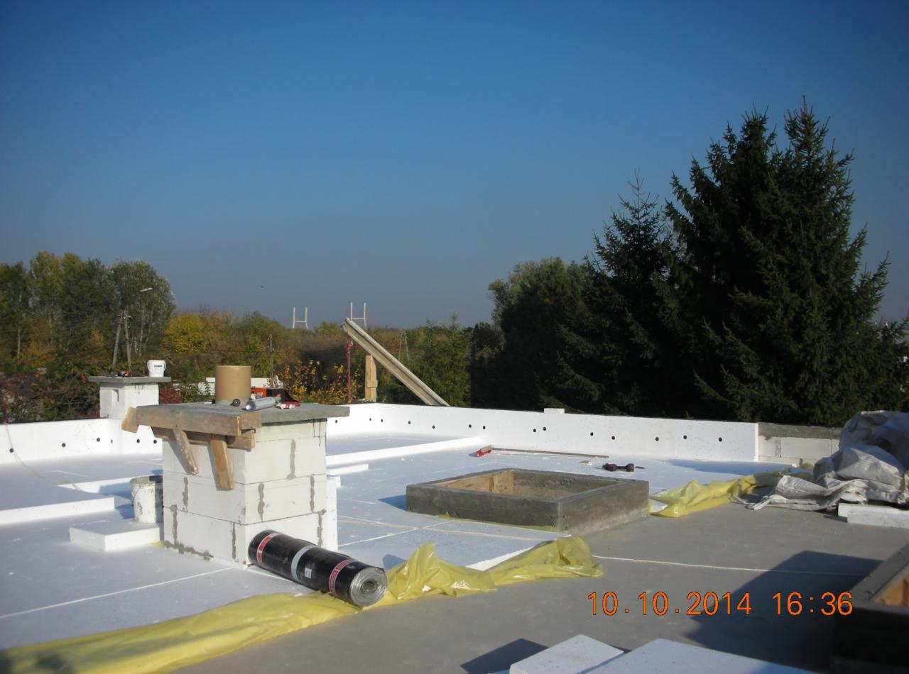 Izolacja płaskiego dachu styropianem i papa termozgrzewalna w rolce, widoczny komin z pustaków i narzędzia, w tle drzewa.