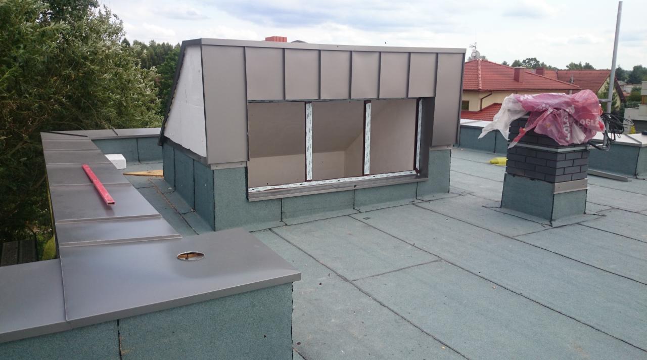 Montaż okna dachowego w obudowie z blachy na pokryciu z papy termozgrzewalnej, widoczny komin w trakcie prac wykończeniowych.