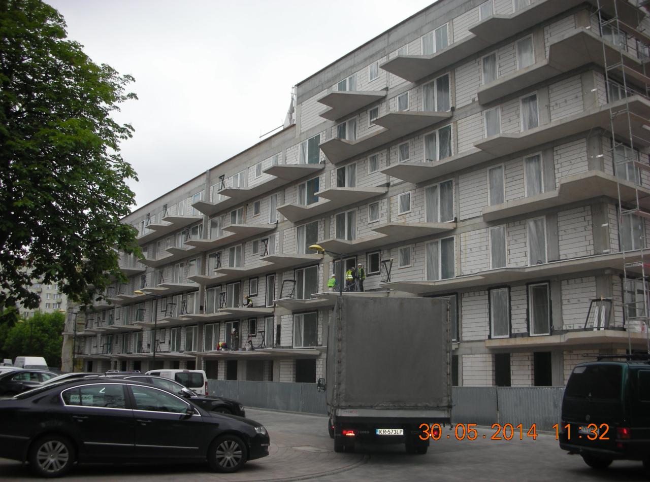 Wielopiętrowy budynek w trakcie budowy z pracownikami na balkonach, widoczny samochód dostawczy i zaparkowane auta.