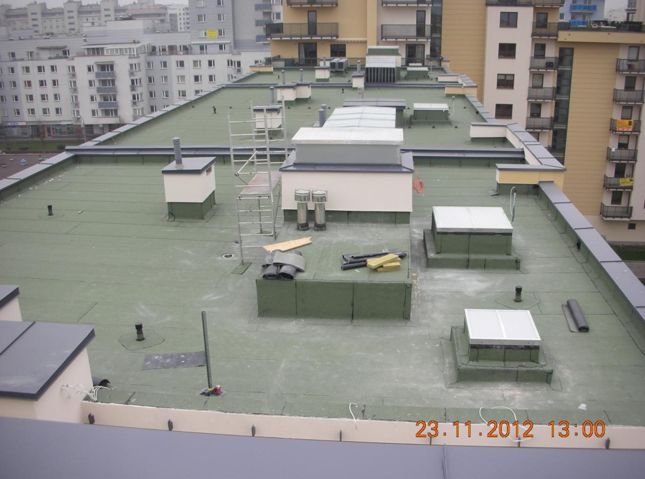 Zielony, płaski dach budynku wielorodzinnego z widocznymi elementami wentylacyjnymi, świetlikami i kominem. Na dachu leżą materiały izolacyjne i rura, a obok stoi drabina.