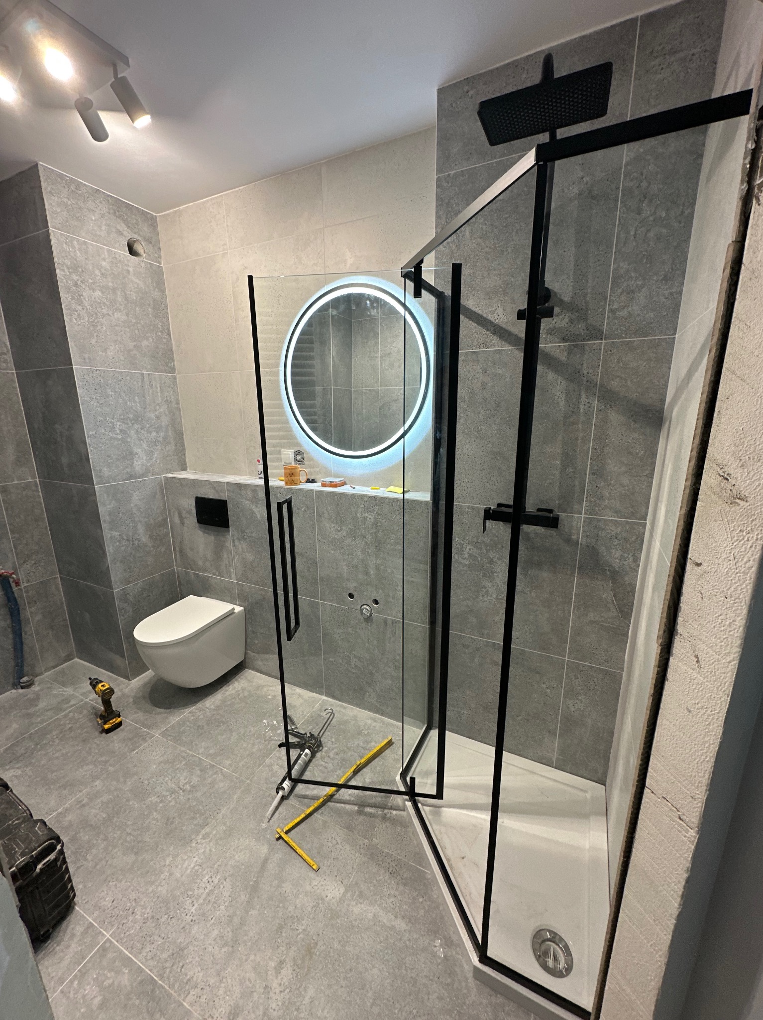 Montaż kabiny prysznicowej z czarnymi profilami w łazience wykończonej szarymi płytkami, widoczne narzędzia i oświetlenie LED.