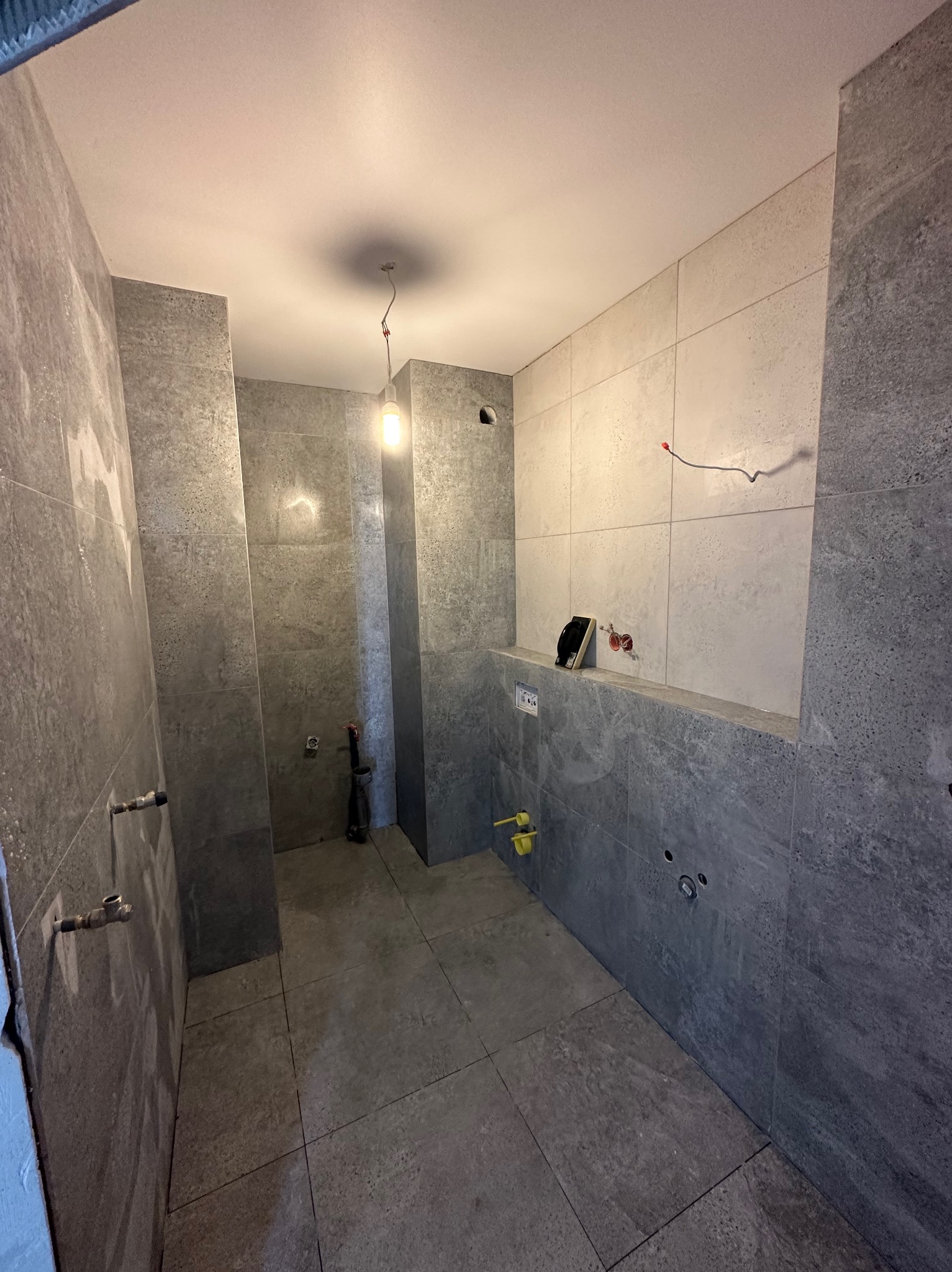 Wnętrze łazienki w trakcie wykańczania, widoczne szare płytki na ścianach i podłodze, wystające przewody elektryczne i rury hydrauliczne, surowa żarówka oświetla pomieszczenie.
