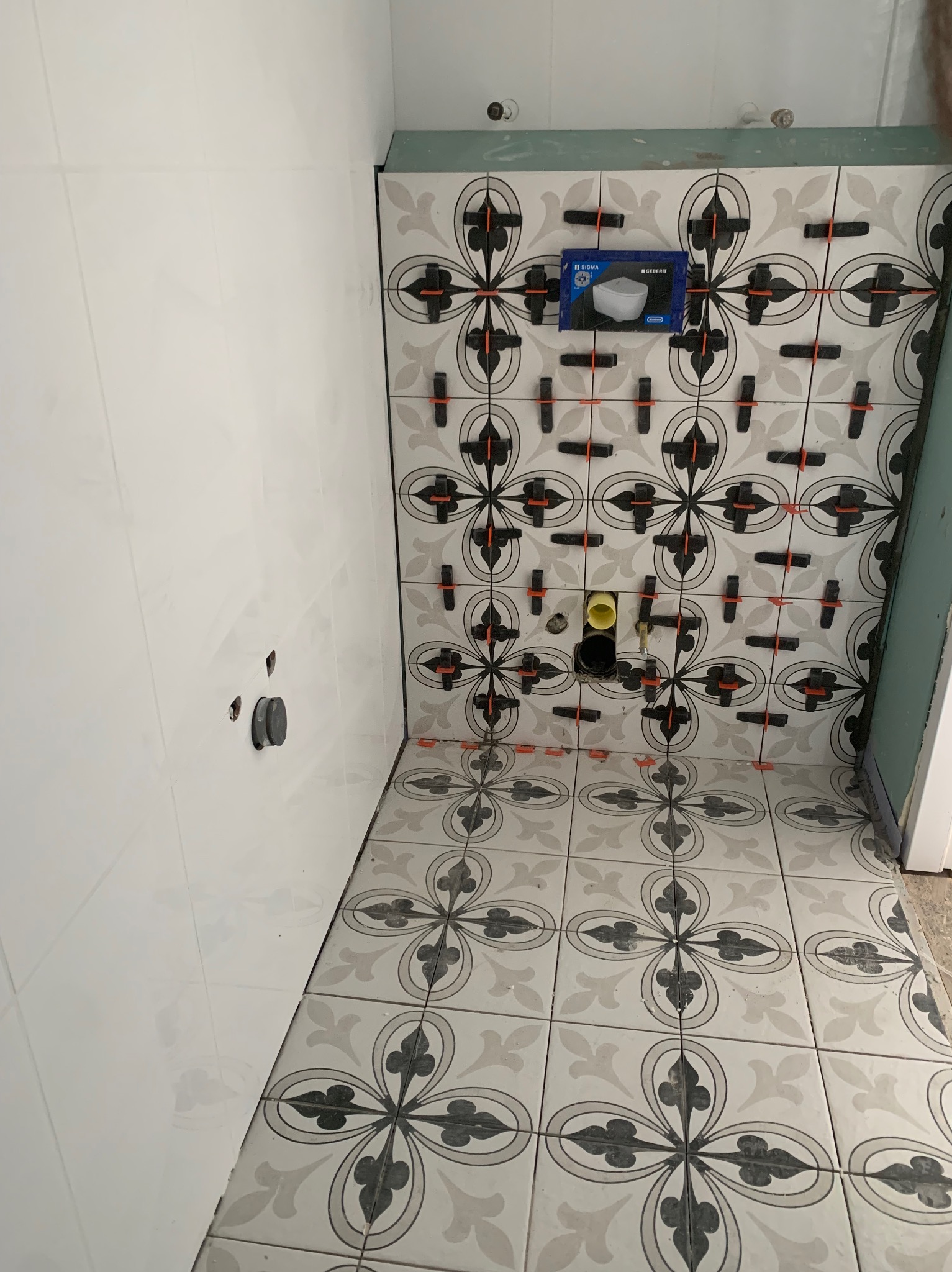 Układanie wzorzystych płytek ceramicznych w łazience, widoczne elementy instalacji i krzyżyki dystansowe, pudełko po stelażu podtynkowym Geberit.