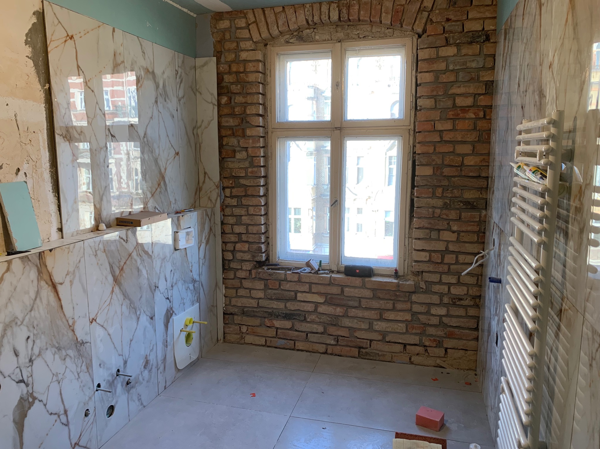 Remont łazienki w trakcie: marmurowe płytki ścienne, ceglana ściana z oknem, grzejnik drabinkowy i elementy instalacji sanitarnej.