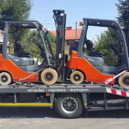 Dwa wózki widłowe Toyota GENEO 15 i Toyota na lawecie transportowej z napisem 'sprzedaż - naprawa - wynajem' na tle zieleni i budynków.