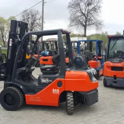 Rząd nowych wózków widłowych Toyota w kolorze pomarańczowo-szarym, stojących na placu pod pochmurnym niebem, z widocznym modelem Tonero 30 SAS na boku.