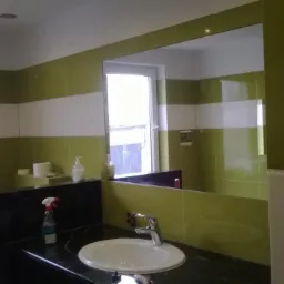 Biała umywalka na tle zielono-białych płytek w łazience z lustrem i oknem. Widoczna armatura i akcesoria łazienkowe.