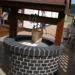 Studnia z czarnej cegły z drewnianym zadaszeniem na kostce brukowej. Wiadro na łańcuchu. Element dekoracyjny ogrodu, wykonany z dbałością o detale.