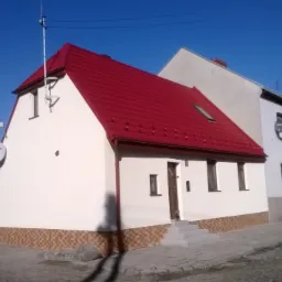 Odnowiony dom z czerwoną blachodachówką i elewacją w odcieniach bieli i beżu. Widoczne okna, drzwi wejściowe i schody. Ujęcie z zewnątrz, w słoneczny dzień.