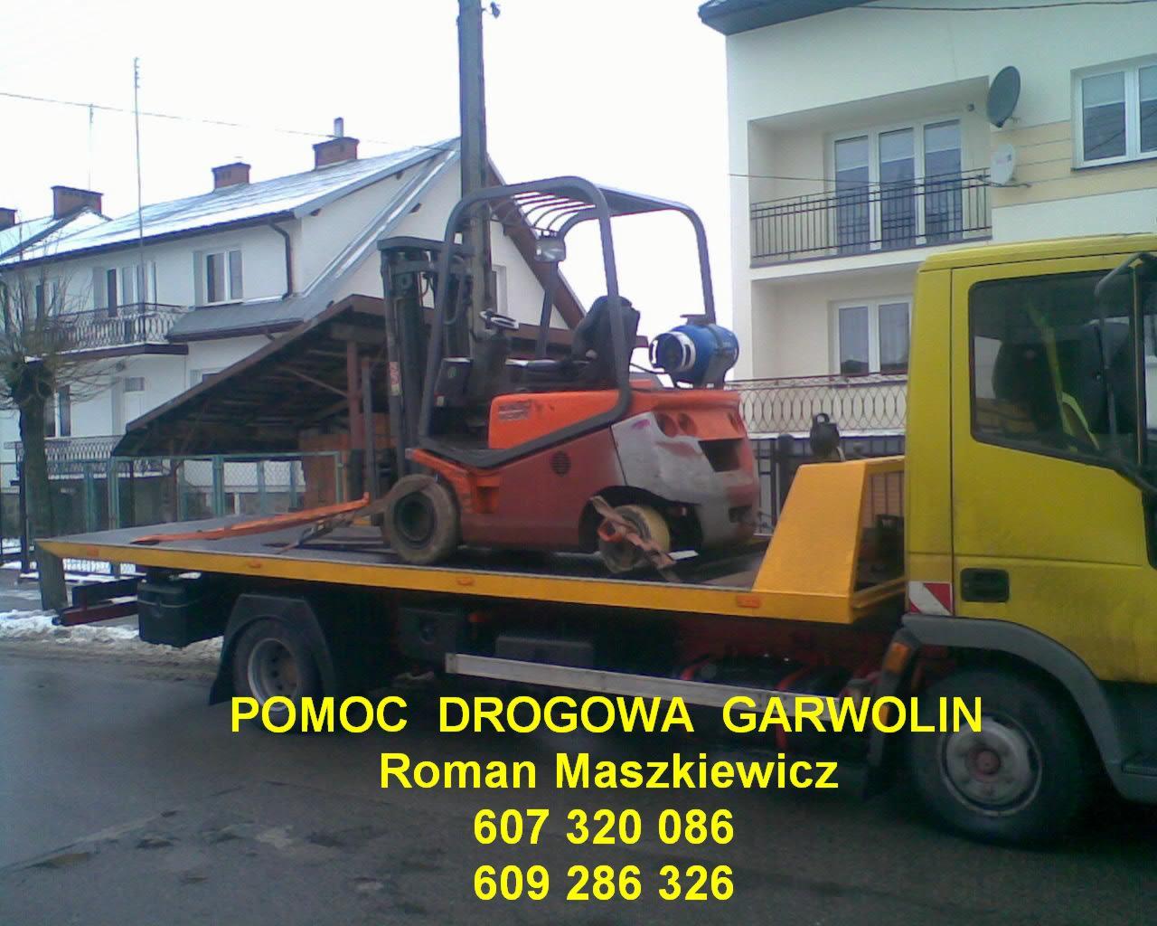 Żółta laweta z pomarańczowym wózkiem widłowym na platformie, widoczne budynki w tle, zdjęcie wykonane w pochmurny dzień.