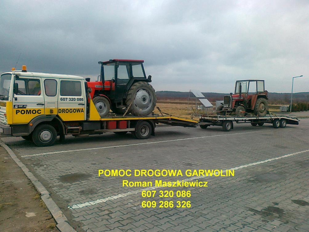 Żółto-biała ciężarówka pomocy drogowej z napisem 'POMOC DROGOWA' przewozi na lawecie czerwony traktor, a za nią druga laweta z kolejnym traktorem na szarym brukowanym placu.