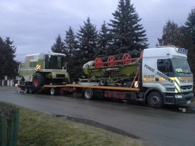 Ciężarówka pomocy drogowej marki Renault przewozi na platformie kombajn zbożowy Claas i jego przystawkę żniwną na tle drzew iglastych.