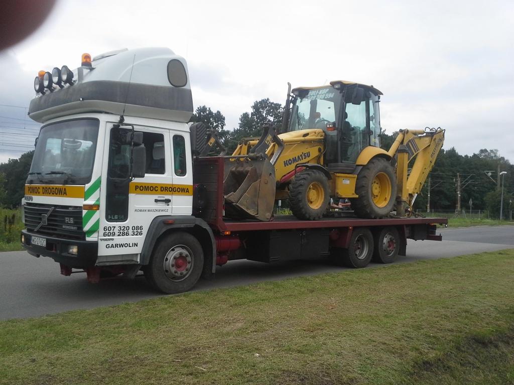 Ciężarówka pomocy drogowej z żółtą koparko-ładowarką Komatsu na platformie, widok z boku na tle zieleni i słupów elektrycznych.