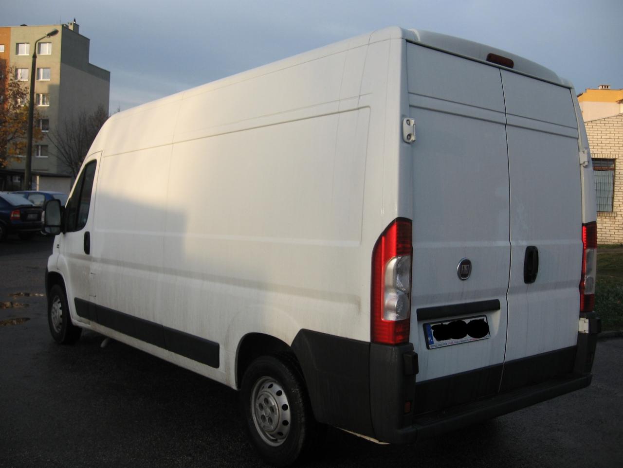 Biały furgon dostawczy Fiat Ducato zaparkowany na mokrym asfalcie, widok z tyłu, z budynkami mieszkalnymi w tle.