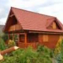 Nowo wybudowany, drewniany dom z bali z czerwonym, dwuspadowym dachem, otoczony zielenią. Widoczny drewniany element dekoracyjny w kształcie lisa.