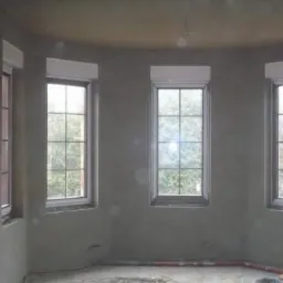 Pokój po nałożeniu szarego tynku maszynowego, z widocznymi czterema oknami z roletami zewnętrznymi, niezabezpieczona żarówka na kablu zwisająca z sufitu.