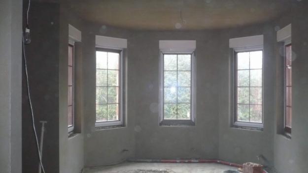 Pokój po nałożeniu szarego tynku maszynowego, z widocznymi czterema oknami z roletami zewnętrznymi, niezabezpieczona żarówka na kablu zwisająca z sufitu.