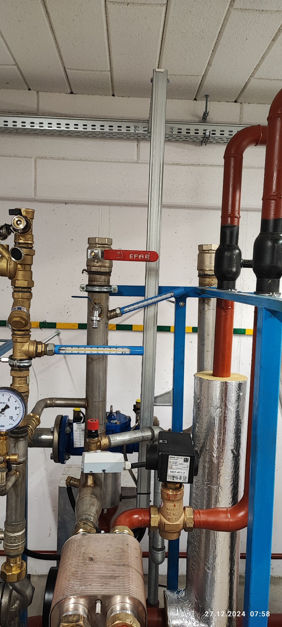 Złożona instalacja grzewcza z rurami miedzianymi, zaworami EFAR i srebrną izolacją termiczną, zamontowana przy białej ścianie z bloków. Widoczny manometr i elementy automatyki.