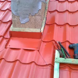 Czerwona blachodachówka na dachu domu, obróbka komina z blachy, narzędzia dekarskie leżą na łacie. Widok z góry na fragment dachu.