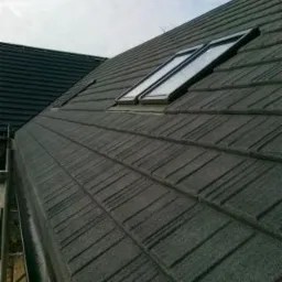 Szary dach z dwoma oknami dachowymi, widok z dołu pod kątem, widoczna rynna i fragment innego dachu w tle.