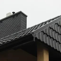 Nowy, czarny dach z błyszczącej dachówki ceramicznej i komin pokryty łupkiem, widoczne obróbki blacharskie i rynna.