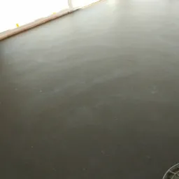 Świeżo wylana, gładka, szara posadzka w jasnym pomieszczeniu, z widocznymi śladami po wygładzaniu i fragmentem okna w tle.