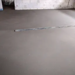 Świeżo wylana, gładka posadzka jastrychowa w szarym odcieniu, z widocznym odpływem liniowym, w surowym wnętrzu budynku z bloczków.