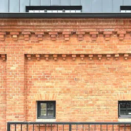 Fragment elewacji z czerwonej cegły z dekoracyjnym fryzem i małymi oknami w czarnych ramach, widok zza czarnej metalowej balustrady.