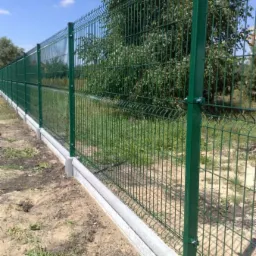 Długi, zielony system ogrodzeniowy z paneli zgrzewanych, zamontowany na betonowej podmurówce, widoczny w słoneczny dzień na tle zieleni.