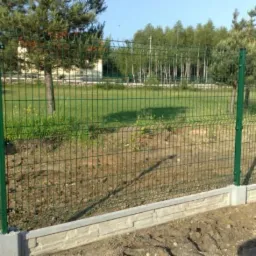 Zielone panele ogrodzeniowe zamontowane na betonowej podmurówce z widocznymi słupkami, w tle trawa i drzewa.